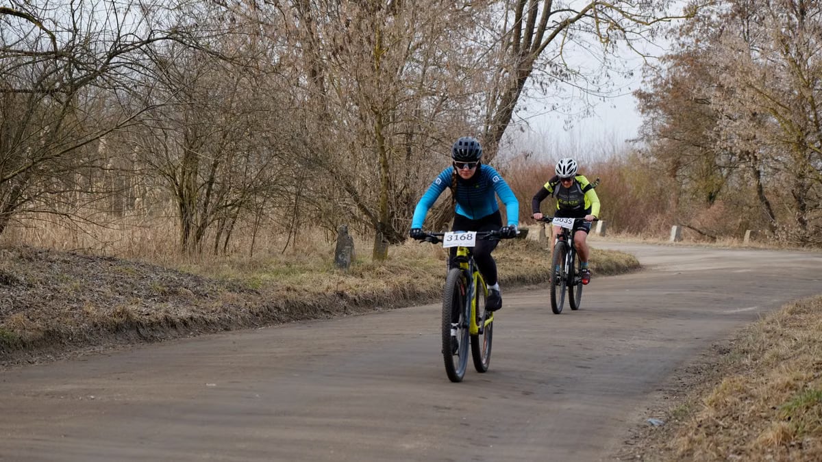 Two cyclists racing on a scenic rural road surrounded by bare trees in early spring.